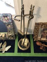Full view of the brass fireplace tool set with poker, brush, shovel, and stand on a display table next to other items.