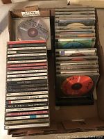 Top-down photo showing a box filled with assorted CDs, stacked vertically and horizontally. Visible titles include Shakiora Oral Fusion volumes, Back to Titanic, Gregorian Chants, Antonio Vivaldi, James Taylor, and others. Some CDs are in jewel cases, others in sleeves.