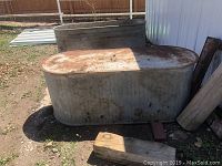 Photo shows two large oval galvanized metal stock tanks outdoors on dirt ground near a shed or barn. Tanks are weathered with rust and discoloration.