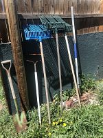 Outdoor garden and stable hand tools including a large spade, garden rake, and three apple pickers with metal baskets supported by wooden handles leaning against a wooden fence.