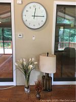 Wide view of wall clock, lamp, and floral arrangements together in room against beige wall near glass doors.