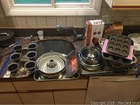 Image showing a variety of baking pans set on a kitchen counter including mini muffin tins, a decorative egg shaped pan, round tube pan, and stacks of pie pans and cookie sheets