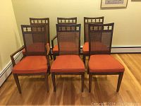 Front view of all six wood frame dining chairs with orange cushions and cane backs, showing two captain's chairs with armrests in front and four armless chairs behind.