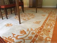 Photo showing cream base area rug with orange decorative motifs under dining table, highlighting wear and pattern details.