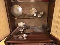 Photo of various silverplate trays, utensils, and glass serving pieces displayed in cabinet with wooden background and cream curtains on either side.