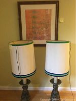 Photo showing both tall table lamps with ceramic bases and off-white shades with green trim in front of the framed print.