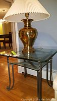 Side table with glass top and shelf, metal frame with curved legs, lamp with reflective brass/gold-tone base and pleated lampshade on top.