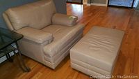 Cream leather armchair paired with matching rolling ottoman on wood floor near a glass side table.