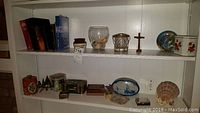 White shelves displaying Bibles and Christianity books, decorative tins, a large scallop seashell, a wire and glass candle holder, souvenir beanpot, religious cross, decorative plates, wooden cigarette box, and assorted small collectibles and keepsake boxes.