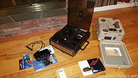 Photo showing Sony reel-to-reel tape recorder in wooden case and Kodak Movie Deck 285 in metal case, placed on wood floor near brick hearth.