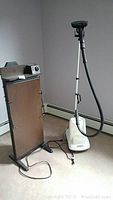 Photo of Conair GS10 fabric steamer and Corby Classic Steam Press pants presser shown side by side on carpeted floor near wall baseboard heater.