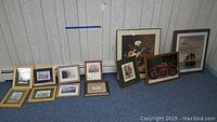 Full view of 12 framed prints arranged on floor against wall showing variety of sizes and subject matter including floral, scenic, and maritime.