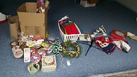 Photo showing assorted Christmas and holiday decor items spread on a carpet including vintage ceramic Christmas tree, artificial wreath, linens, tins, and gift bags.