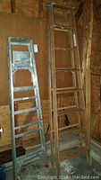 Side by side view of 6 ft aluminum ladder and 8 ft wooden ladder leaning against pegboard wall in garage with visible signs of wear