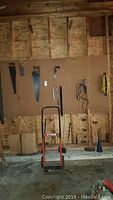 Photo showing multiple saws hanging on pegboard, a hand truck in front, axes, and other tools on wooden wall in garage.