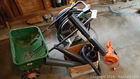 Photo of green seed spreader, orange leaf blower, and black hose on portable spool in garage setting