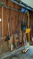 Photo showing a variety of yard tools hung on a garage pegboard and wall including shovels, pitchfork, pickaxe, rakes, and snow shovel.