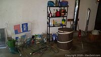 Lot contents against a white wall including a green garden hose coiled on the concrete floor with spray nozzle, assorted containers and bottles on a black metal shelving unit, and a brown pop-up canvas barrel.