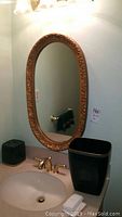 Photo of oval ornate framed mirror above bathroom sink with matching black wastebasket and tissue holder featuring gold trim visible