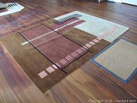 Area rug laid flat on a wooden floor, showing full rug with geometric pattern in browns, pink, cream, black, and beige colors.