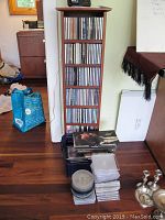 Tall wooden CD stand filled with multiple rows of CDs. Several box sets and additional CDs are stacked on the floor in front of the stand.