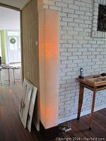 Full view of the tall fabric shade floor lamp standing against a white brick wall and wood panel wall, showing lamp illuminated with warm light.