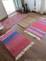 Photo showing all four area rugs laid out on a hardwood floor near a doorway. The rugs show various colors including reddish tones and multicolor stripes with fringe.