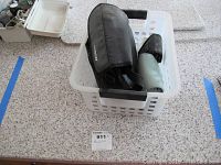 Three binoculars in cases inside a white laundry basket on a speckled floor.