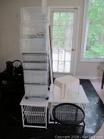 Plastic storage unit on wheels with drawers, plastic stool atop small white metal stands with wire baskets, near a glass door and window.