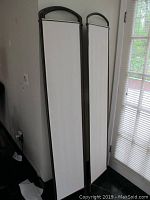 Three metal frame room dividers with white cloth panels, seen partially folded leaning against a wall.