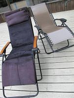 Two folding lounge chairs on a wooden deck, one black padded with wooden armrests, the other beige mesh with black armrests.