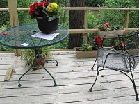 Two wrought iron outdoor tables with round mesh tops and two matching wrought iron mesh chairs on a wooden deck area surrounded by plants and trees.