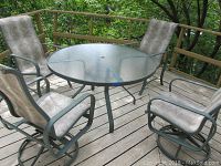 Wide view of the round glass top patio table with four matching chairs on wooden deck with surrounding greenery.