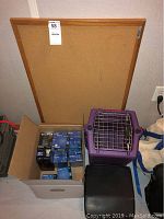 Photo of a cork board against the wall, a box containing Sony and Fujifilm data tapes, a purple pet carrier with metal front door, and a black typewriter case.
