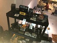 Two black plastic shelves holding various vintage and modern cameras, camera bags below and accessories scattered. Cameras include vintage Kodak and Hawk-Eye models alongside newer Panasonic and Nikon cameras.