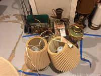 Photo showing three vintage lamps including one with a clear glass base and fabric shades, with cords and plugs visible, placed on a concrete floor with other items nearby.