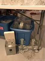 Photo of cut glass pitchers, covered bowls, and various glassware arranged in blue bins and under a folding table.