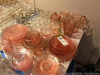 Photo showing a collection of rose colored Depression glass including pitchers, plates, and serving pieces, alongside several sets of clear drinking glasses with silver or gold rims.