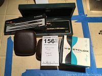 Cross pen and keychain in black Cross-branded box with Parker pen set and two vintage Westclox travel clocks in brown cases and original boxes, all arranged on floor marked with blue tape.