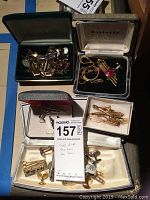 Photo of cuff links and tie clips in two open boxes, showing gold-tone and silver-tone accessories with some featuring decorative stones or engravings.