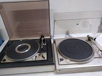 Two vintage Dual turntables side by side, one with a tinted dust cover and the other with a clear dust cover. Shows general wear and damage to the cover of the Model 1225 unit.