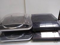 Top view of four stacked turntables, two with silver bases and clear dust covers, two with black bases including Optimus and Denon brands.