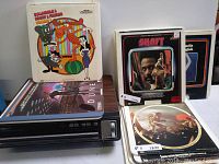 Photo showing RCA SelectaVision VideoDisc Player with several video disc sleeves stacked on top and around device. Titles visible include Hanna-Barbera cartoons, Shaft, and Dune.