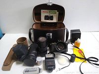 Photo showing Pentax MV camera body, lenses, leather carrying case, flash unit, manual, and various camera accessories laid out on a white surface.