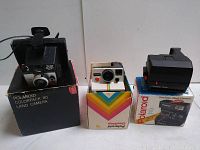 Three vintage Polaroid cameras displayed with their original boxes, positioned side by side including Colorpack 80, OneStep, and OneStep Flash Camera.