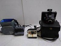 Photo showing both vintage Polaroid cameras and their respective carrying cases in a side-by-side view on a white surface.
