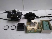 Two vintage film cameras with accessories, including a black Nikon EM camera and a silver Sears camera with strap, metal rings and camera cases