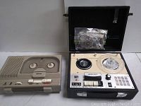 Both vintage reel to reel tape recorders side by side on a flat surface. The Granada unit is open with a plastic reel on the right spindle and a plastic bag with cables stored inside the case. The Fajiya unit is closed with its transparent cover showing reels.