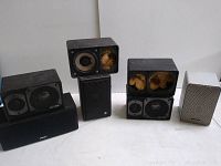 Photo shows seven vintage speakers of various sizes and conditions on white floor against white wall, some with exposed speaker components and yellow foam padding inside.