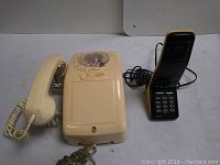 View of 2 vintage telephones on white surface, showing rotary dial phone and cordless-style phone side by side.